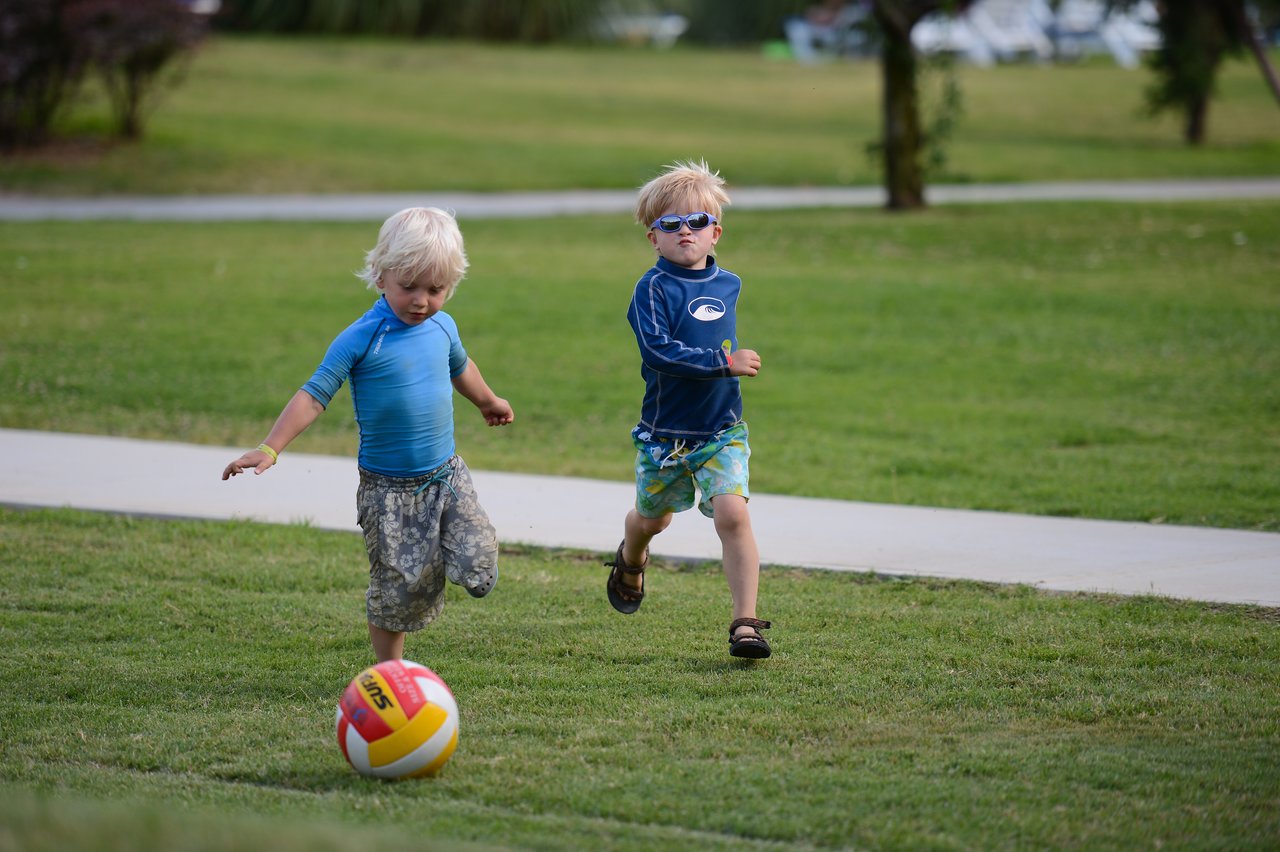 Two young children play on a grassy field, one kicking a volleyball while the other runs behind.