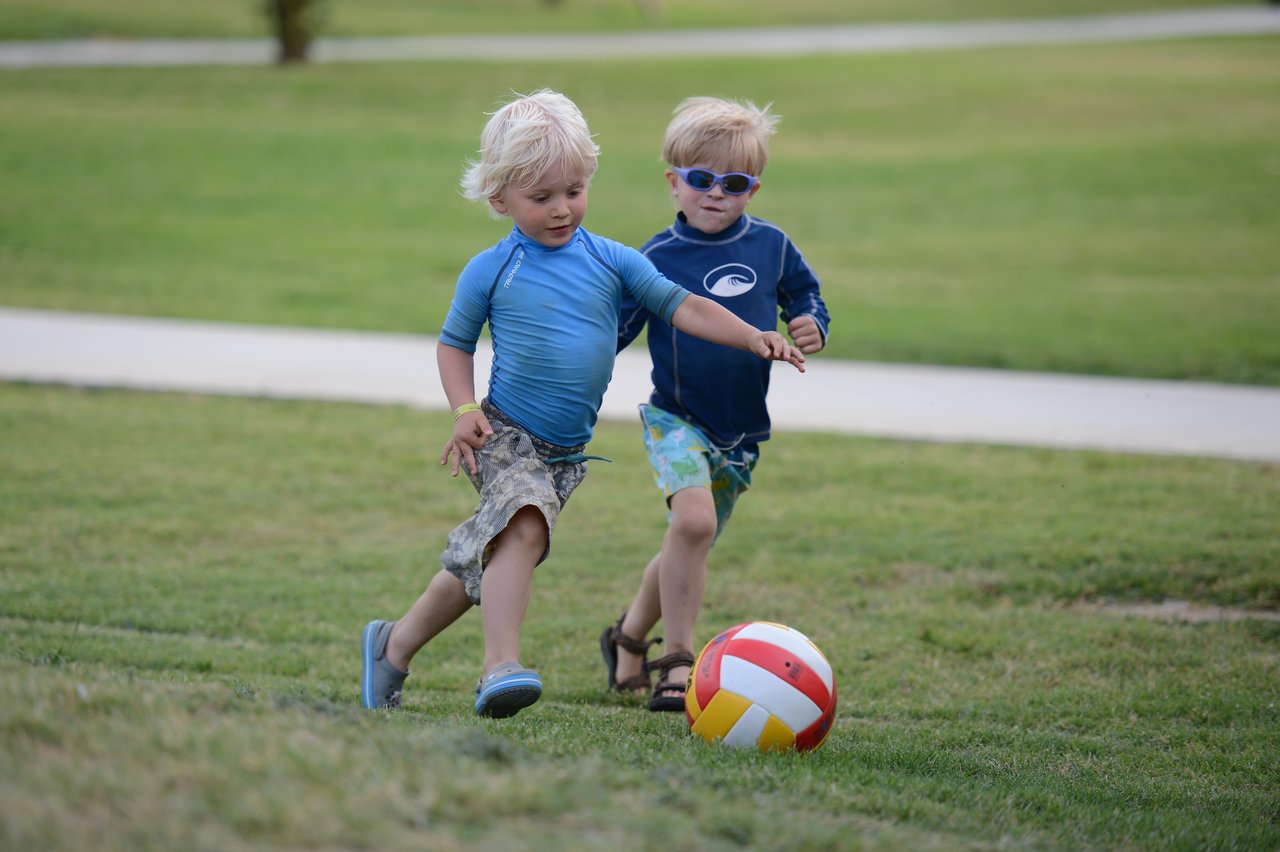 Two young children play soccer on a grassy field, chasing a red, yellow, and white ball.