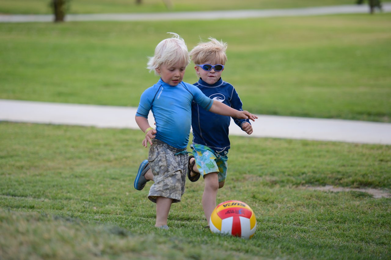 Two young children play soccer on a grassy field, chasing a colorful ball while running barefoot.