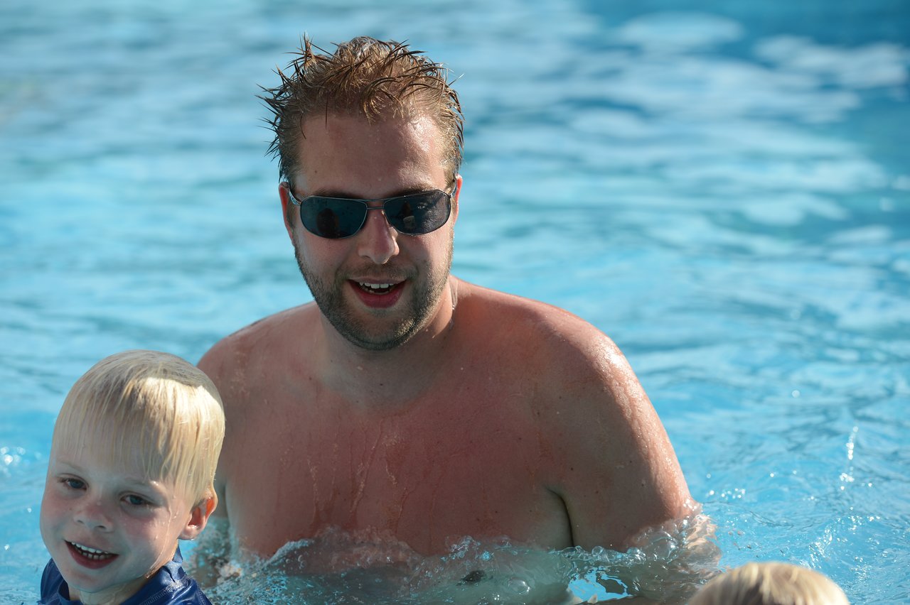 A man wearing sunglasses is in a swimming pool with two children, smiling and enjoying the water.