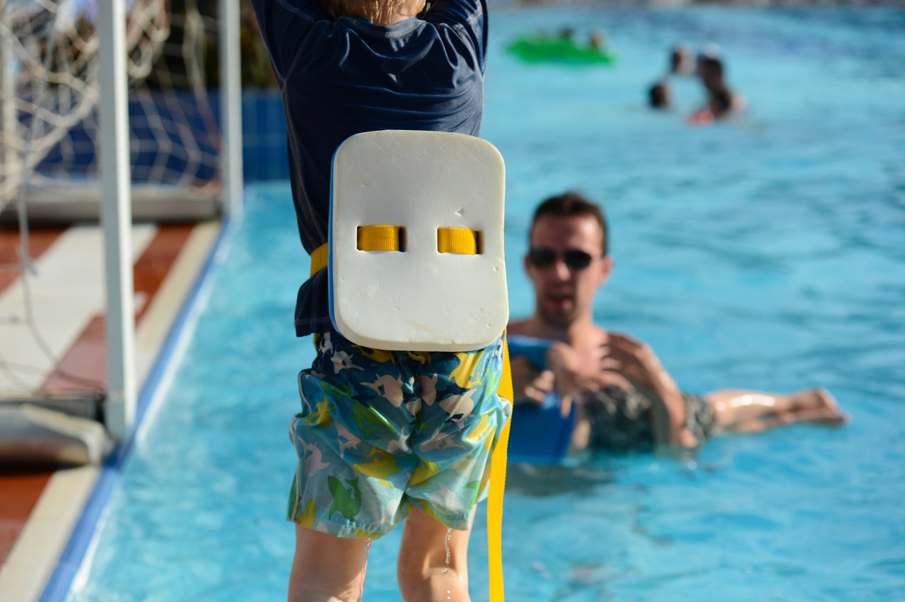 A child wearing a flotation device prepares to jump into a swimming pool while an adult watches nearby.