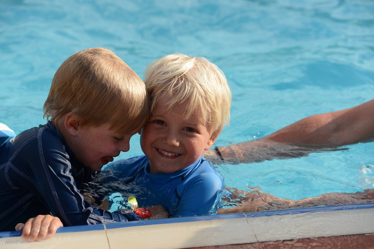 Two young children in swimwear play and smile while leaning on the edge of a swimming pool.