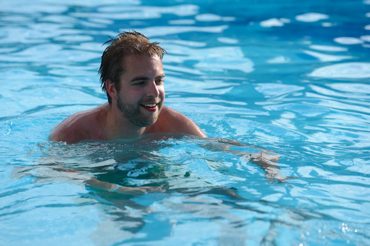 A man with wet hair swims in a clear blue pool, smiling.