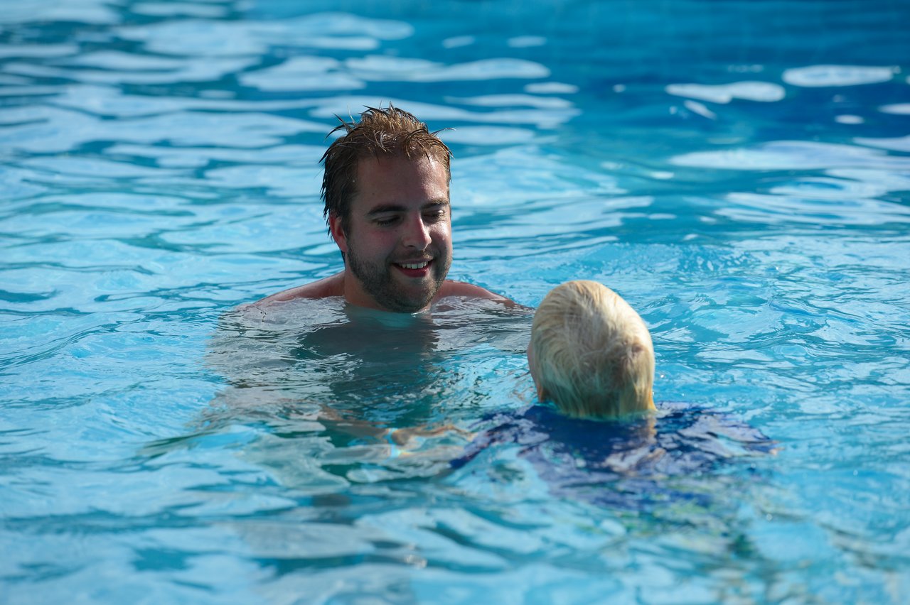 A man and a child swim in a pool, with the man smiling at the child.