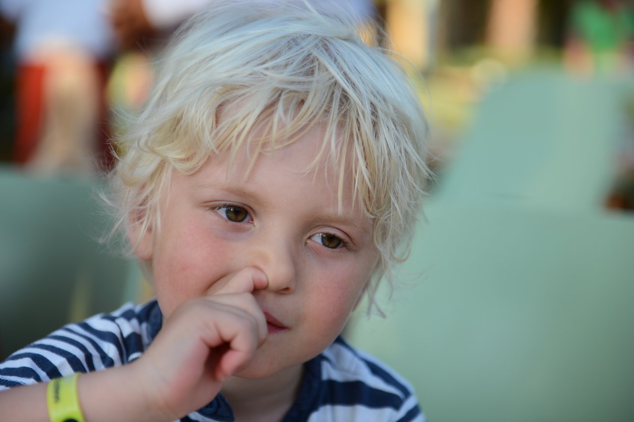 A young child with blonde hair picks their nose while looking slightly away from the camera.
