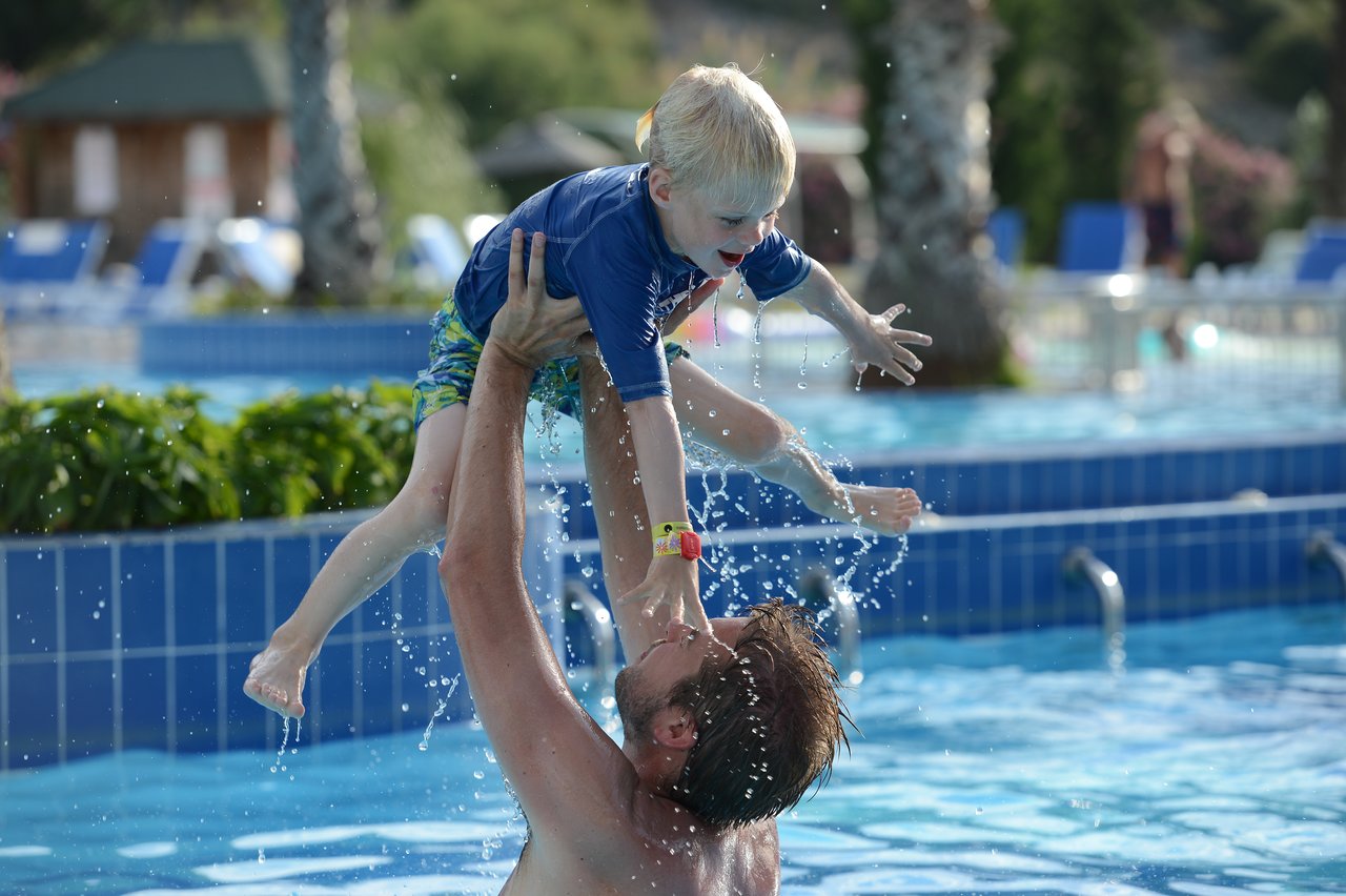 An adult lifts a smiling child out of the water in a swimming pool, splashing water around them.