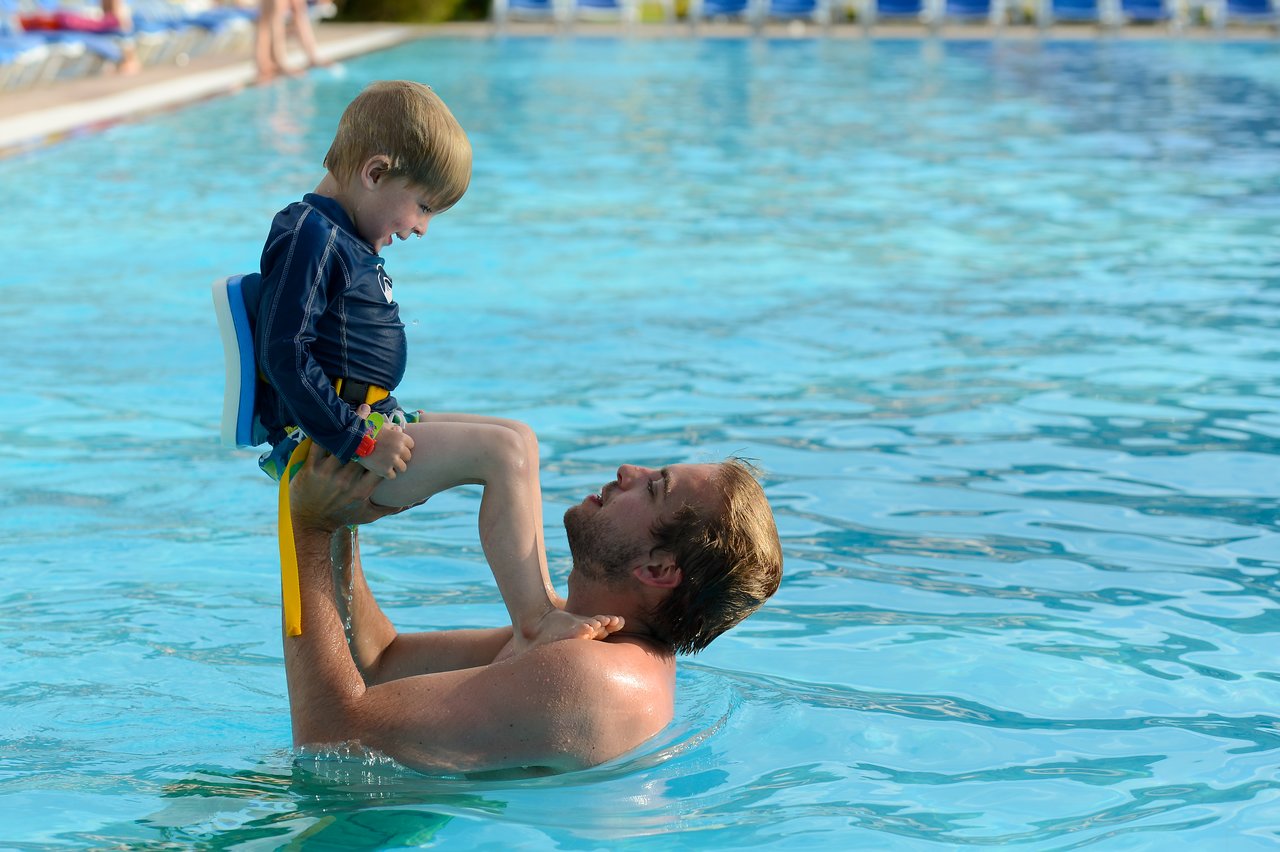 A man in a pool lifts a young child wearing a flotation device and swimwear.