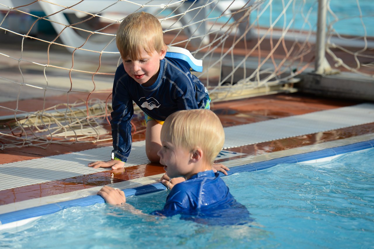 A child in the pool looks up at another child kneeling by the edge, both wearing swim shirts.