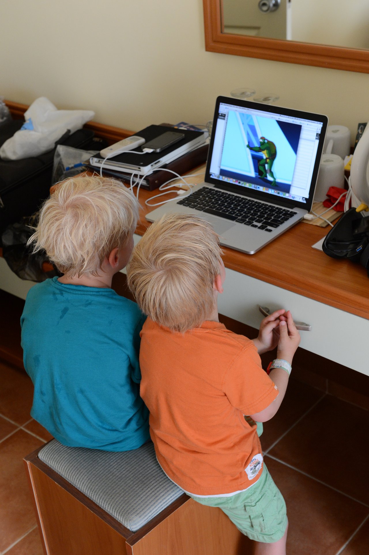 Two children with wet hair sit on a stool, watching an animated show on a laptop screen.