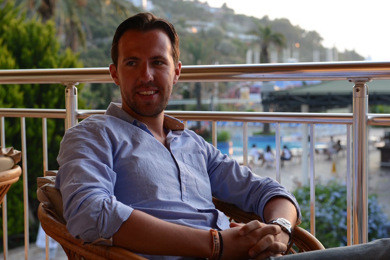 A man in a blue shirt sits on a balcony, smiling at the camera with a relaxed posture.