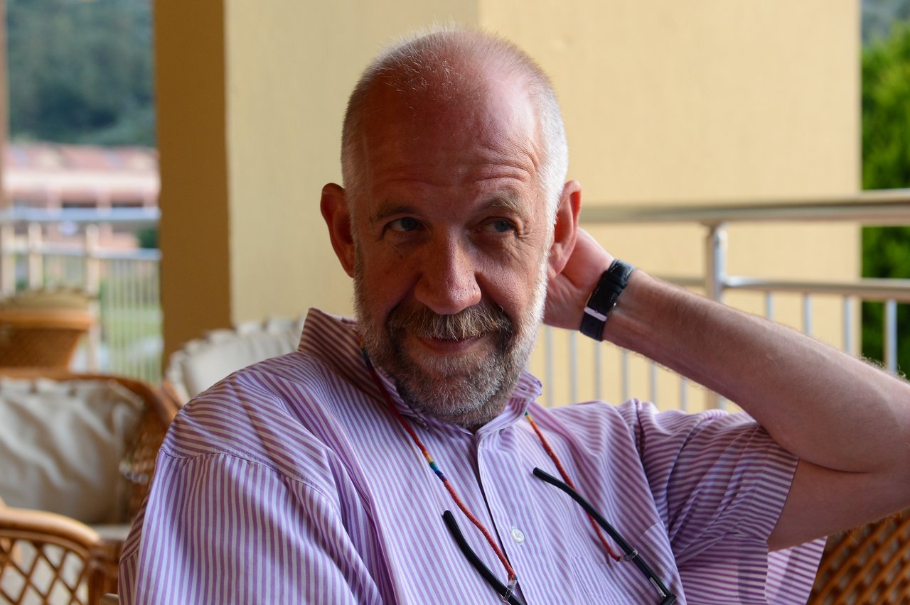 A man in a striped shirt sits outdoors, resting his hand on his neck and smiling slightly.