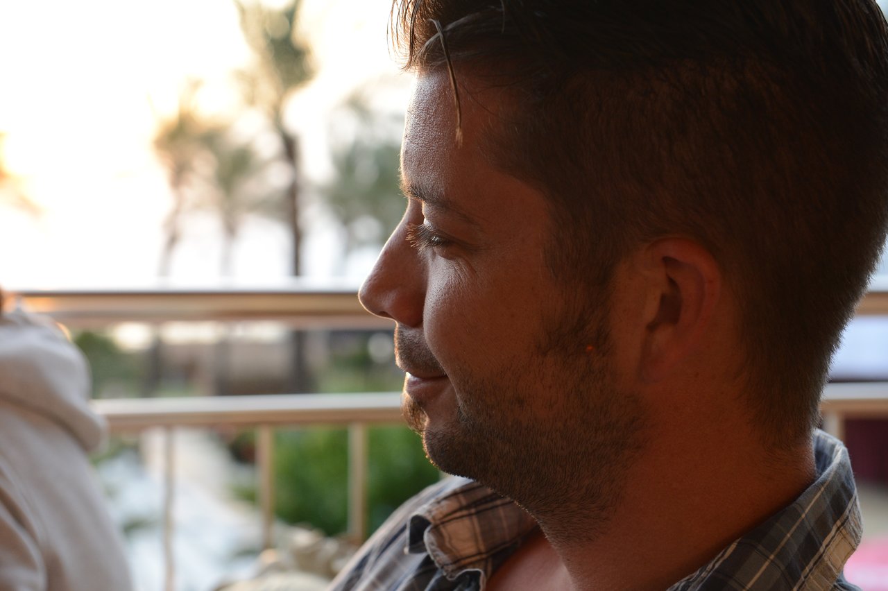 A man with short hair and a beard sits outdoors, looking to the side with a relaxed expression.