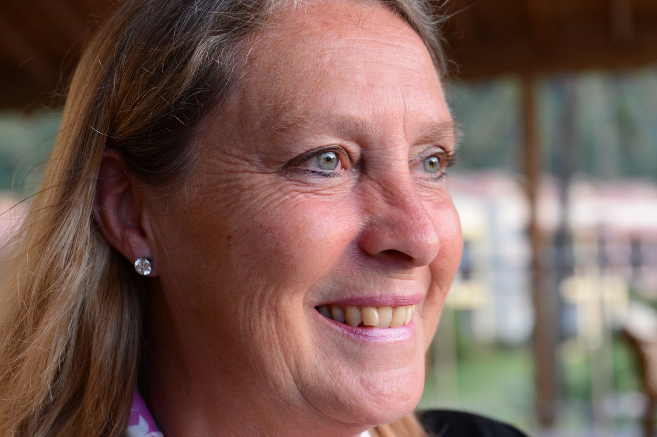 A woman with long hair and earrings smiles while looking off to the side in an outdoor setting.