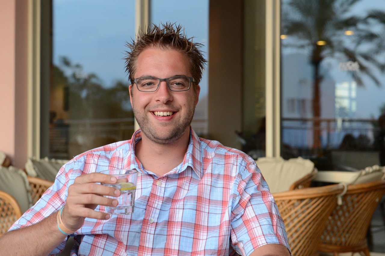 A smiling man in glasses and a plaid shirt holds a glass with a lemon slice at an outdoor café.