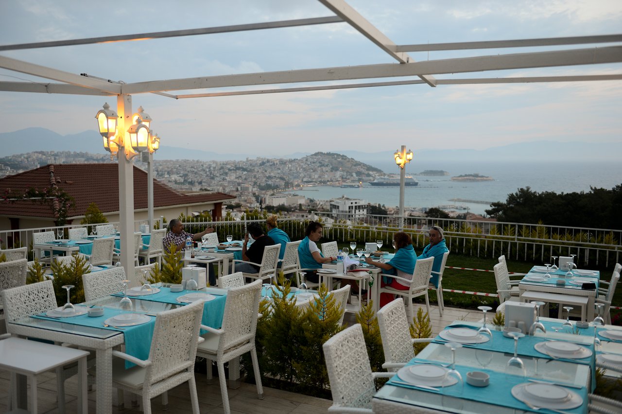 People dining at an outdoor restaurant with a view of the sea and city in the background.