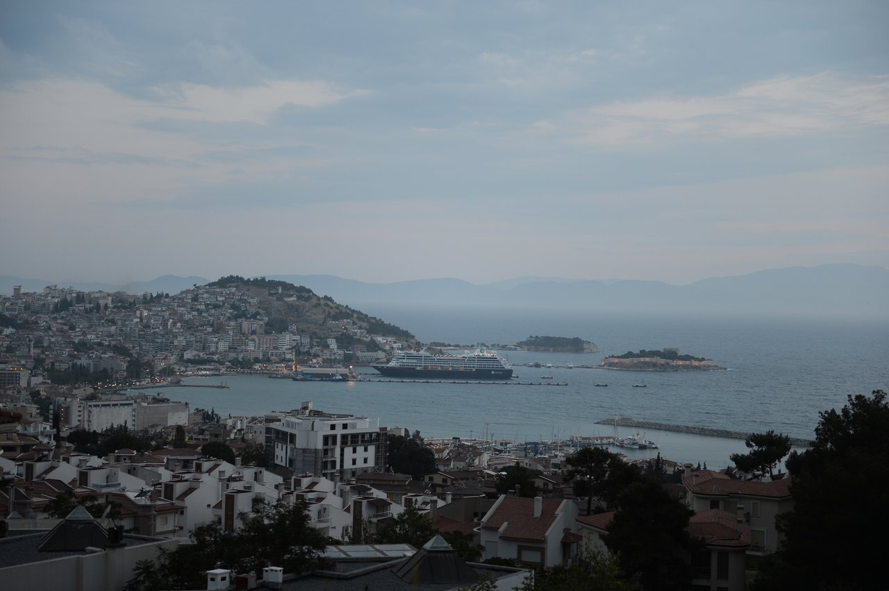 A large cruise ship is docked near a coastal city with hills, buildings, and small islands in the background.