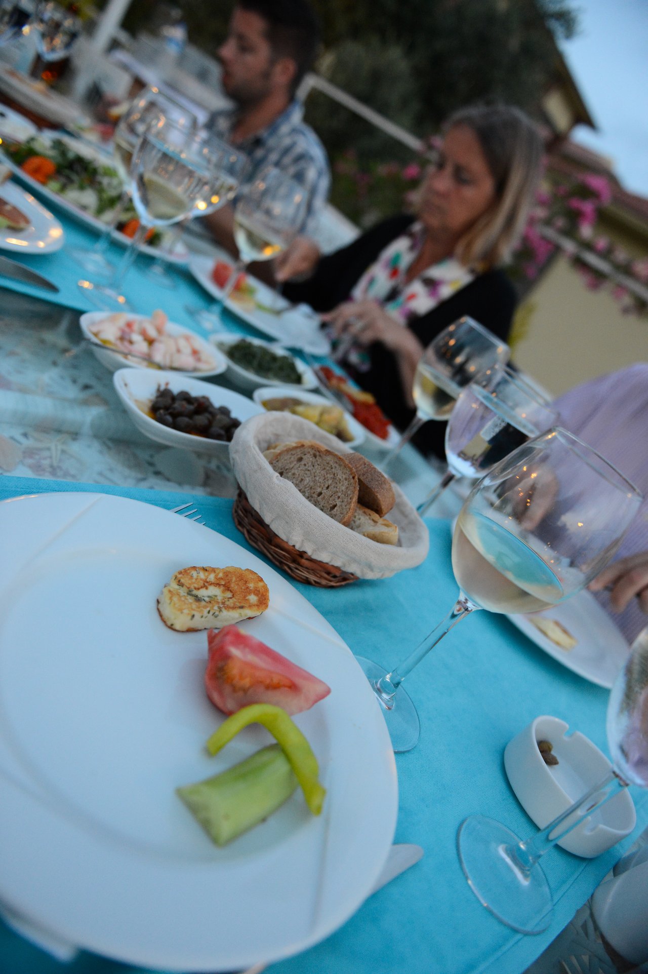 People dining at an outdoor table with plates of food, bread, and glasses of white wine.