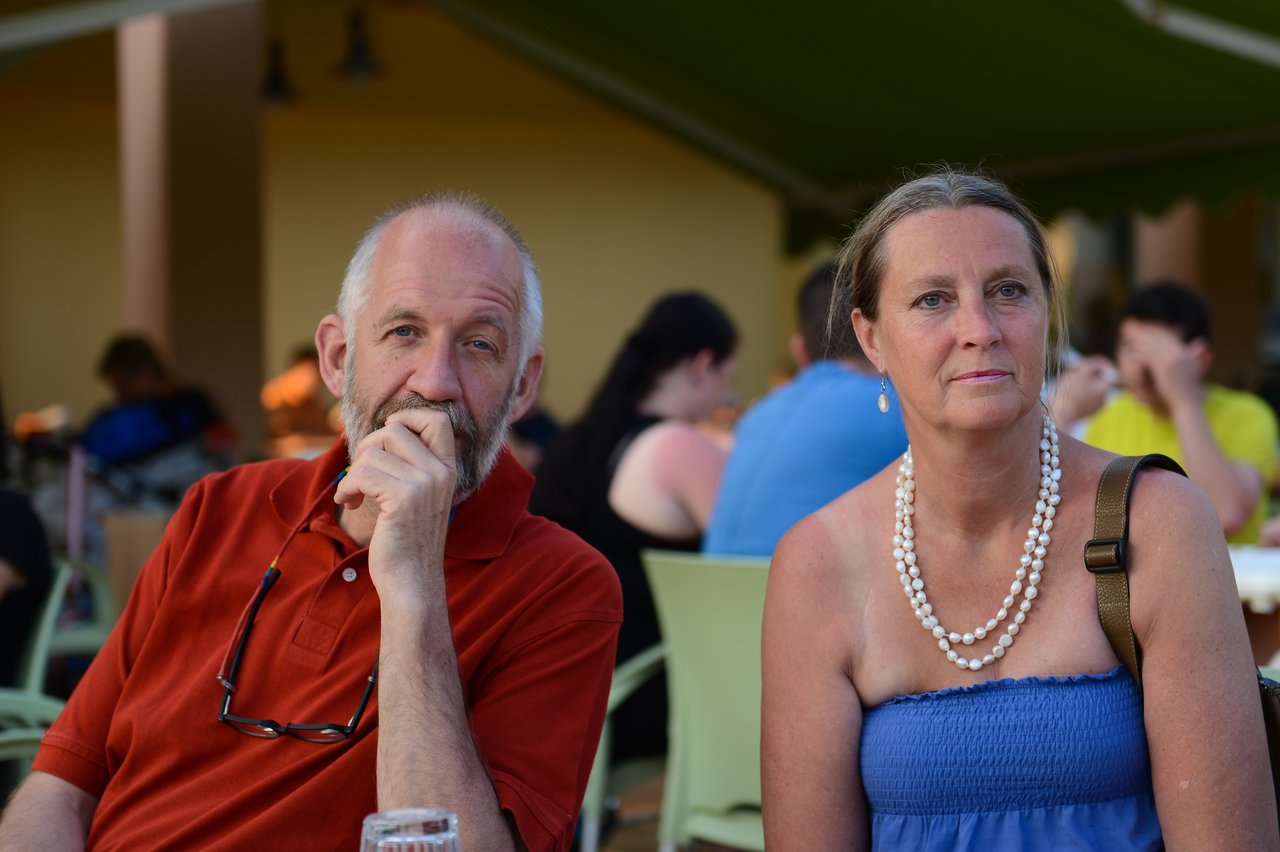 A man and a woman sit at an outdoor café, looking in different directions with neutral expressions.