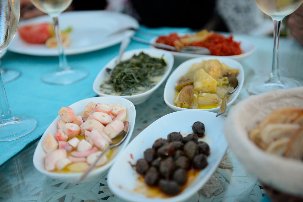 A table with small plates of Turkish appetizers, including olives, seafood, and vegetables, alongside glasses of white wine.