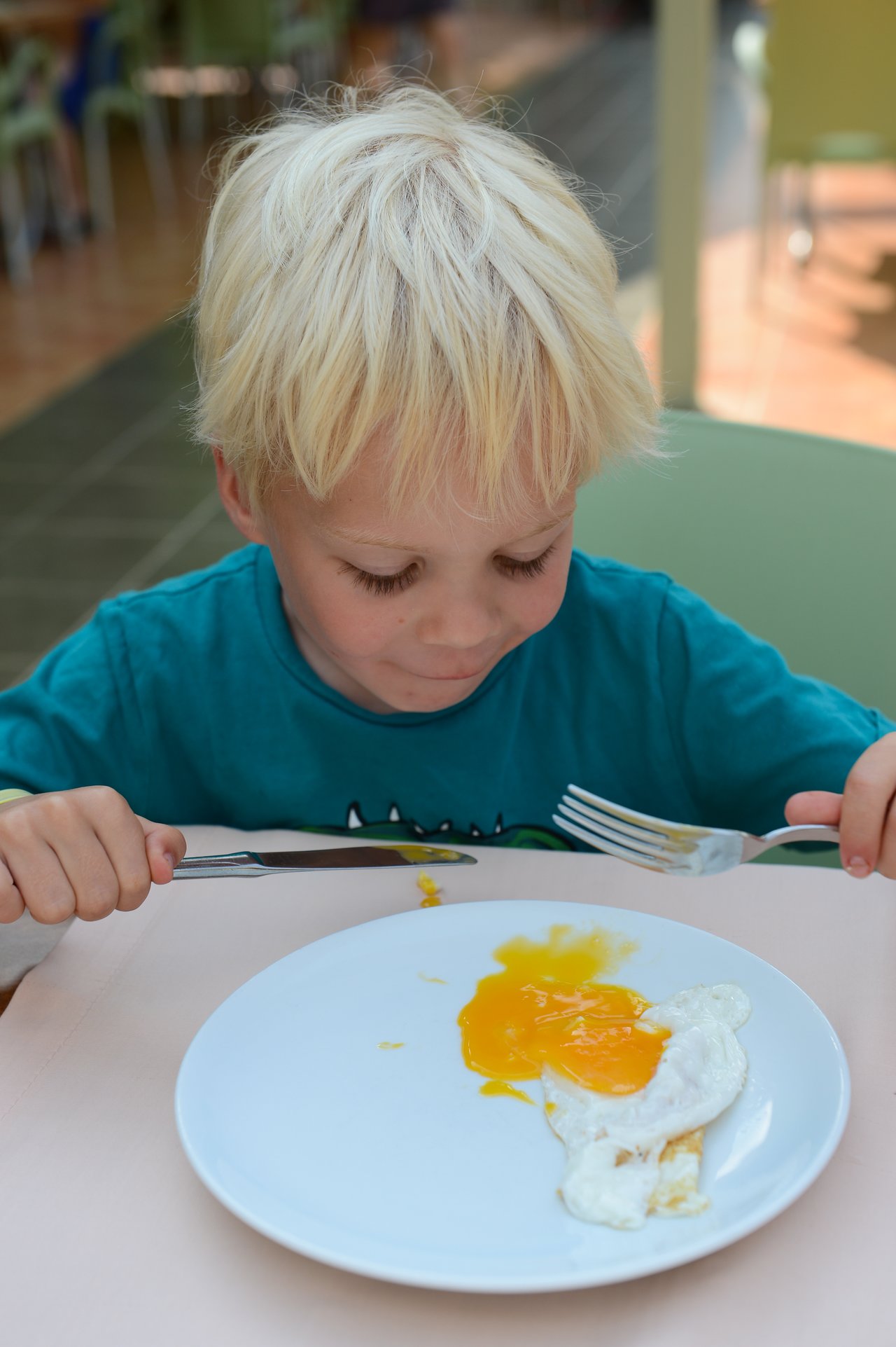 A child in a blue shirt holds a fork and knife, looking at a plate with a broken fried egg.