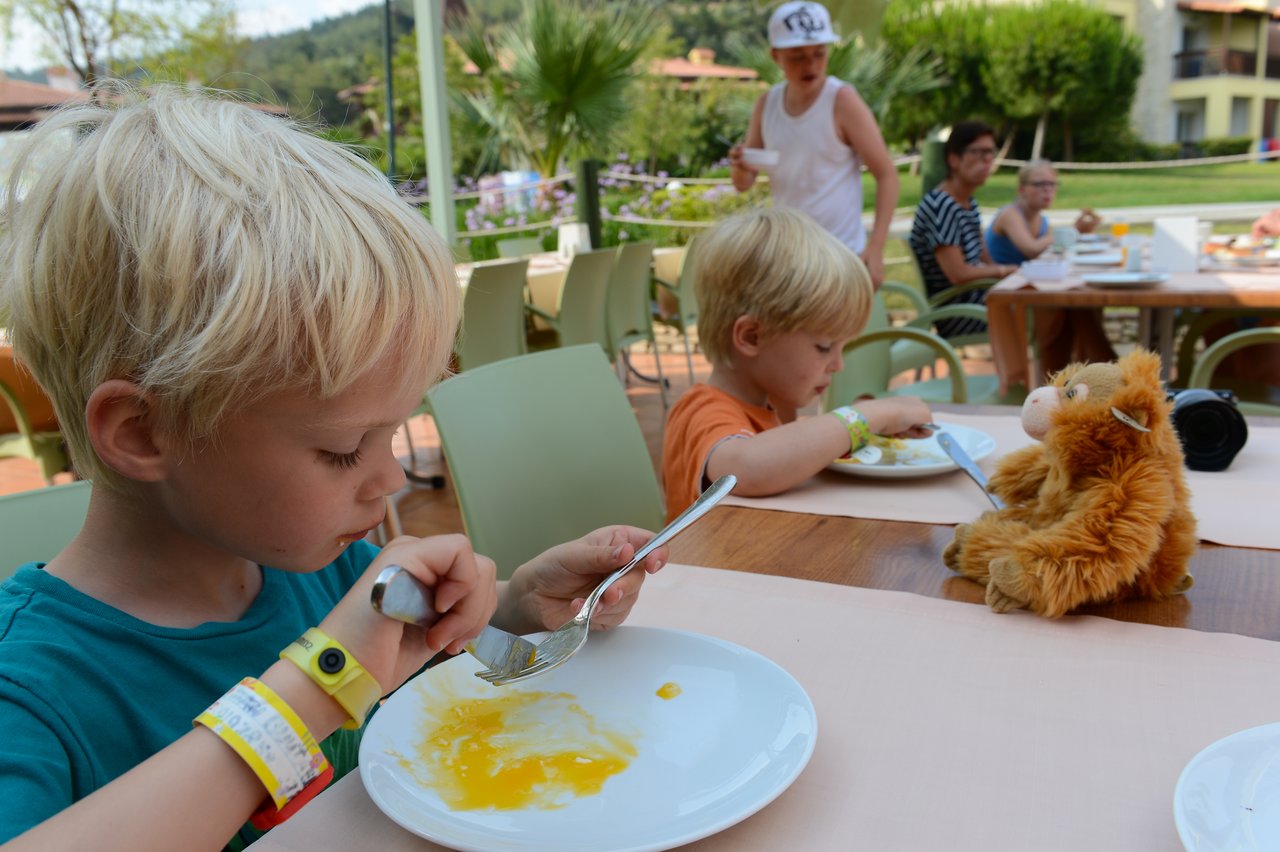 Two young children eat at an outdoor restaurant, with a stuffed monkey toy sitting on the table nearby.
