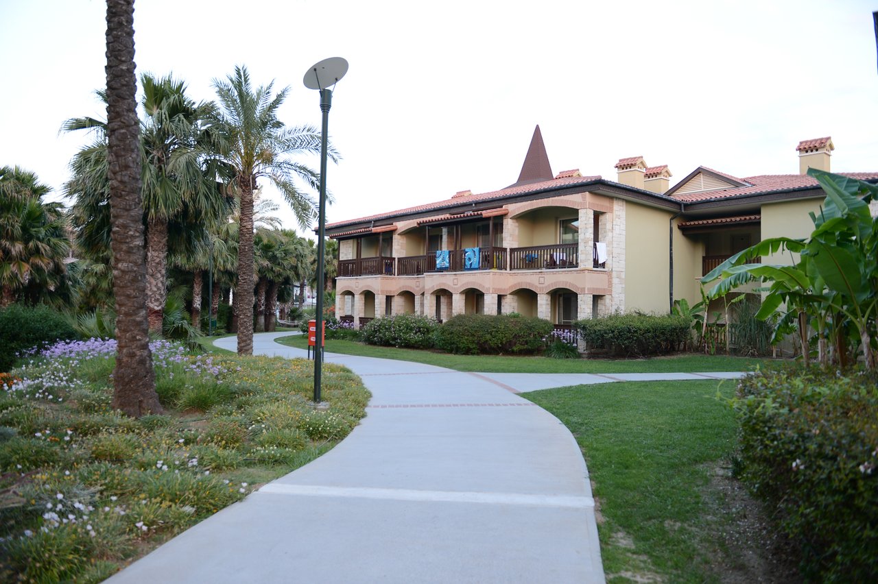 A paved walkway leads to a two-story building with balconies, surrounded by palm trees and green landscaping.