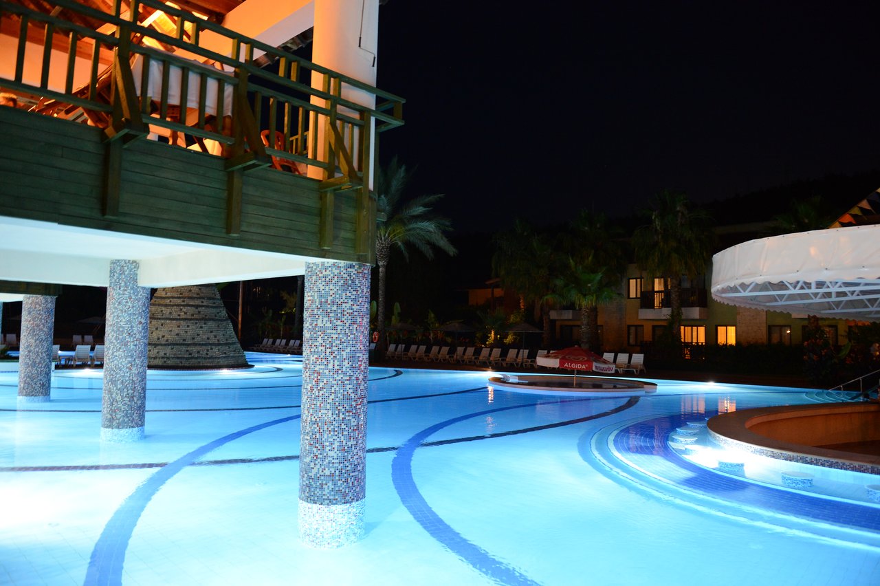 A well-lit swimming pool at night with a wooden balcony and surrounding resort buildings in the background.