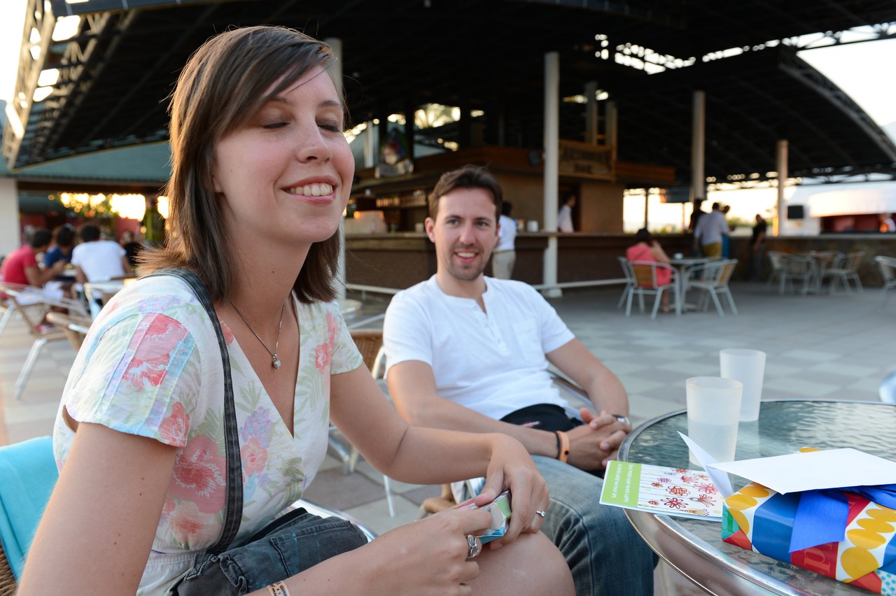 A woman smiles with her eyes closed while sitting at an outdoor table, with a man smiling behind her.