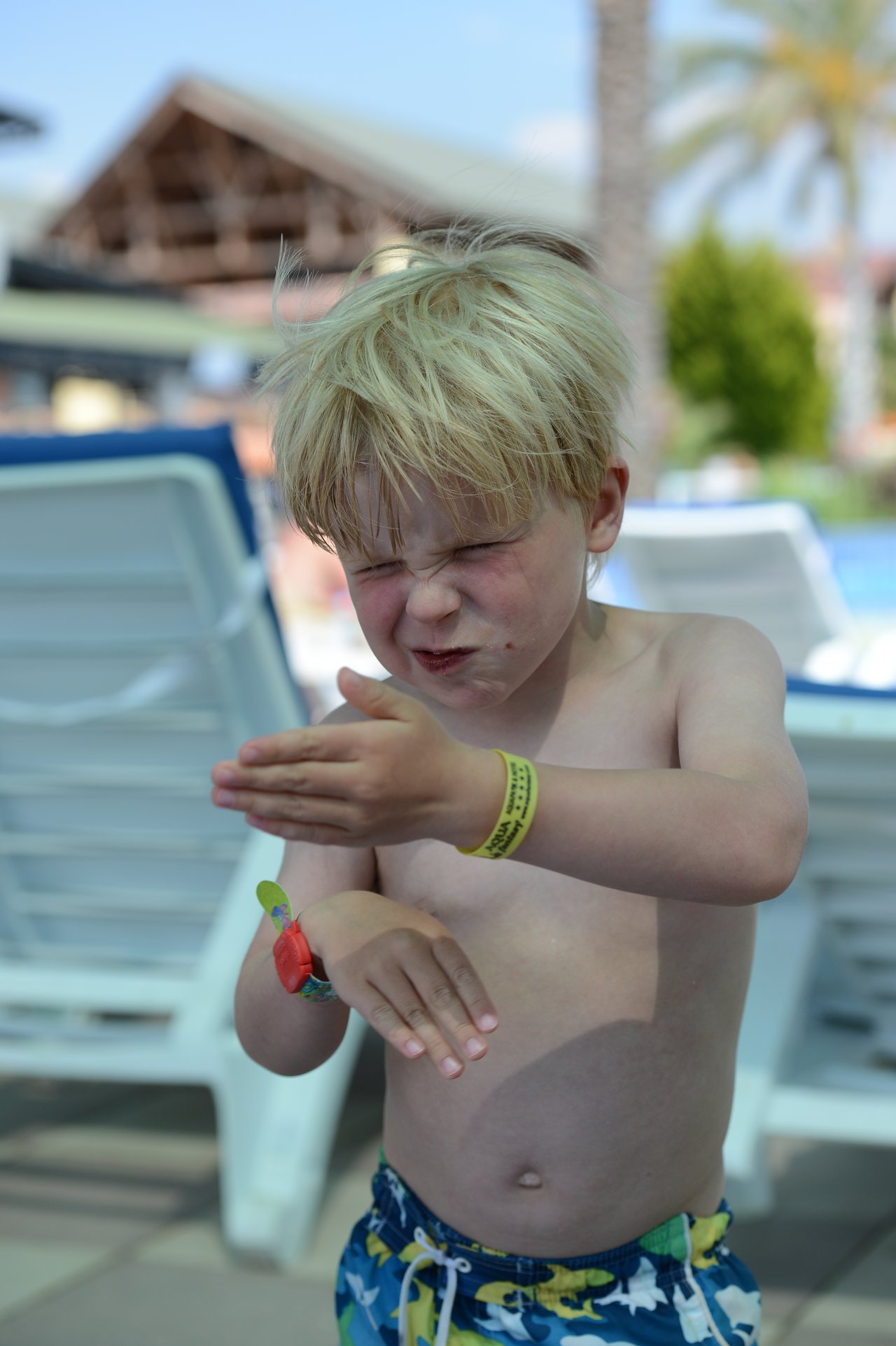 A young boy in swim trunks squints and rubs his hands together near a poolside lounge chair.