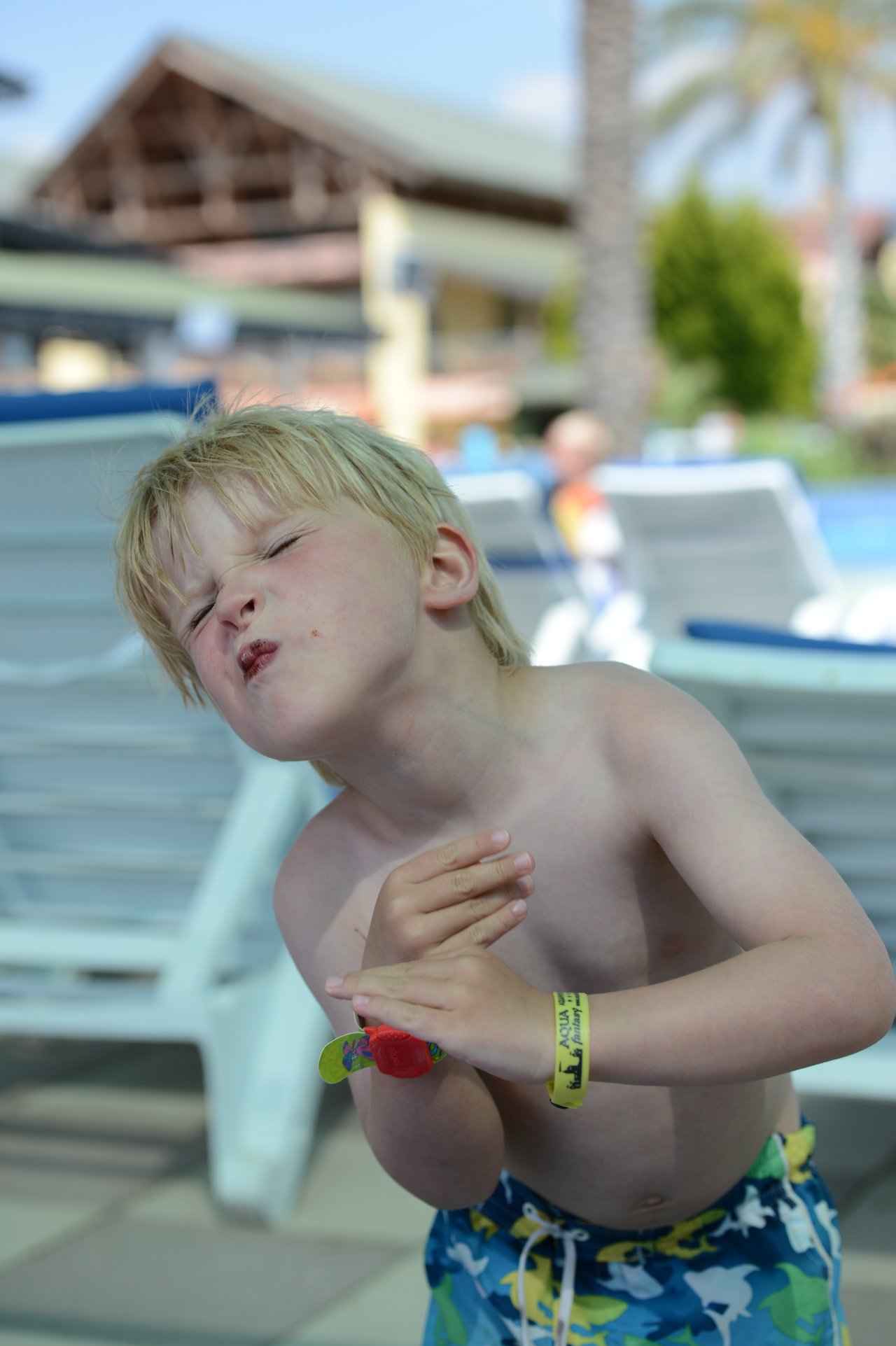 A young boy in swim trunks makes a playful face and gesture near a poolside lounge chair.