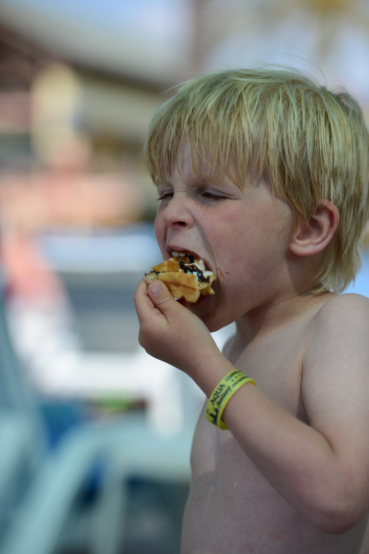A young child with blond hair takes a big bite of a sandwich while wearing a yellow wristband.