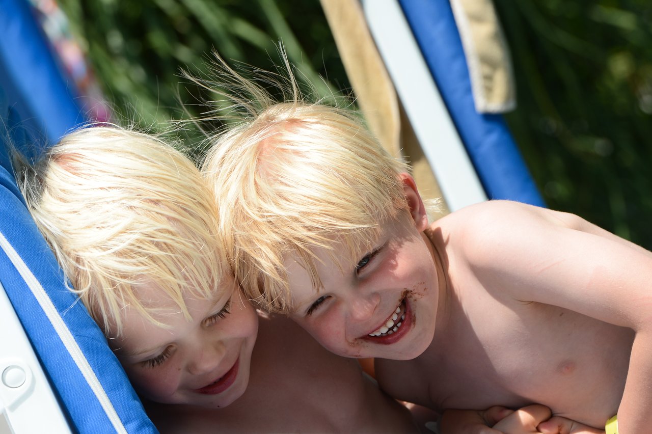 Two young children with blonde hair smile and lean close together on a blue lounge chair outdoors.