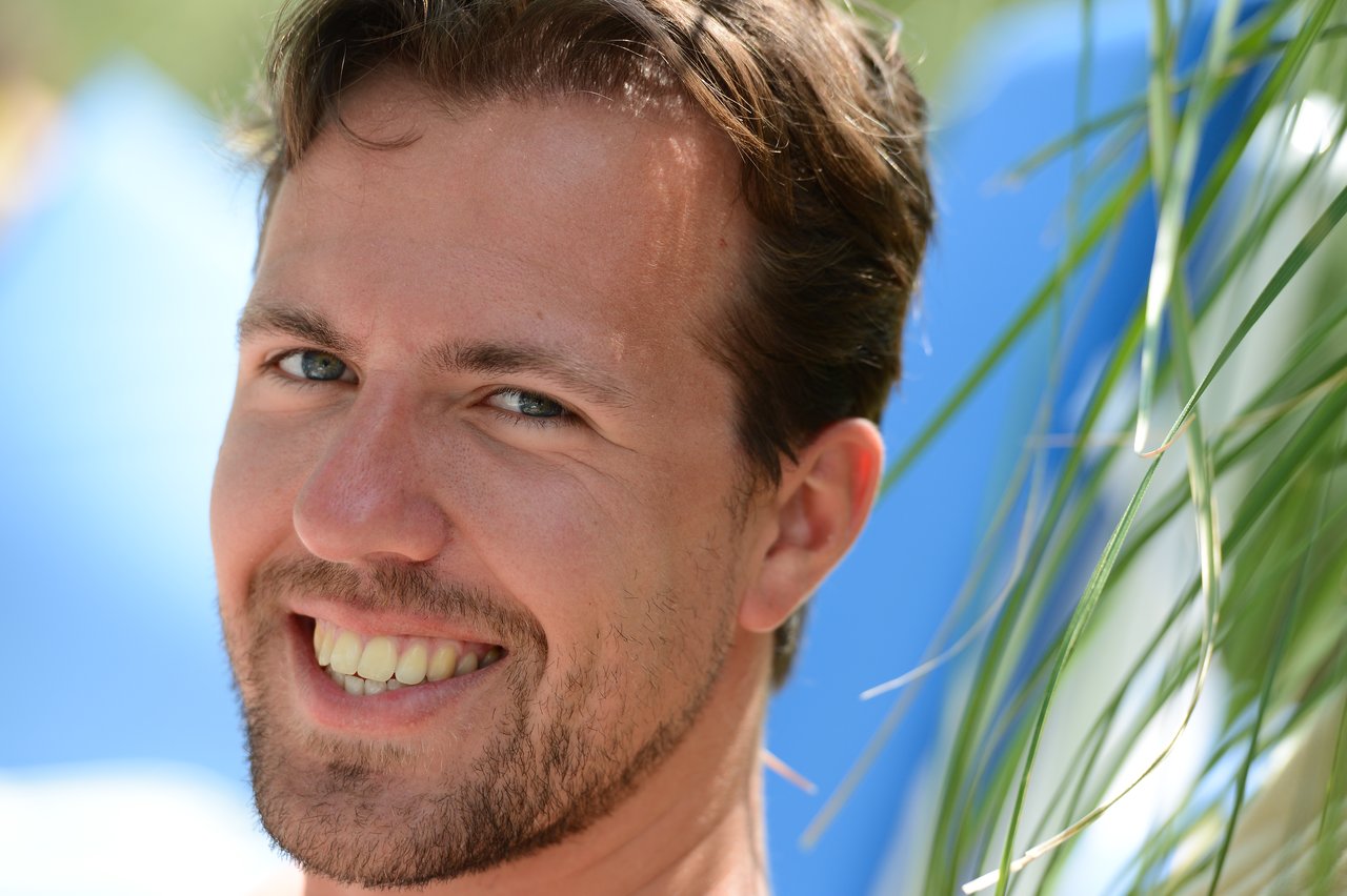 A man with short brown hair and a beard smiles at the camera, with blurred greenery in the foreground.