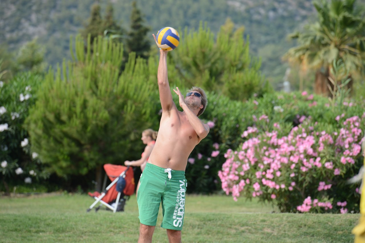 A man in green shorts prepares to hit a volleyball outdoors, with greenery and flowers in the background.