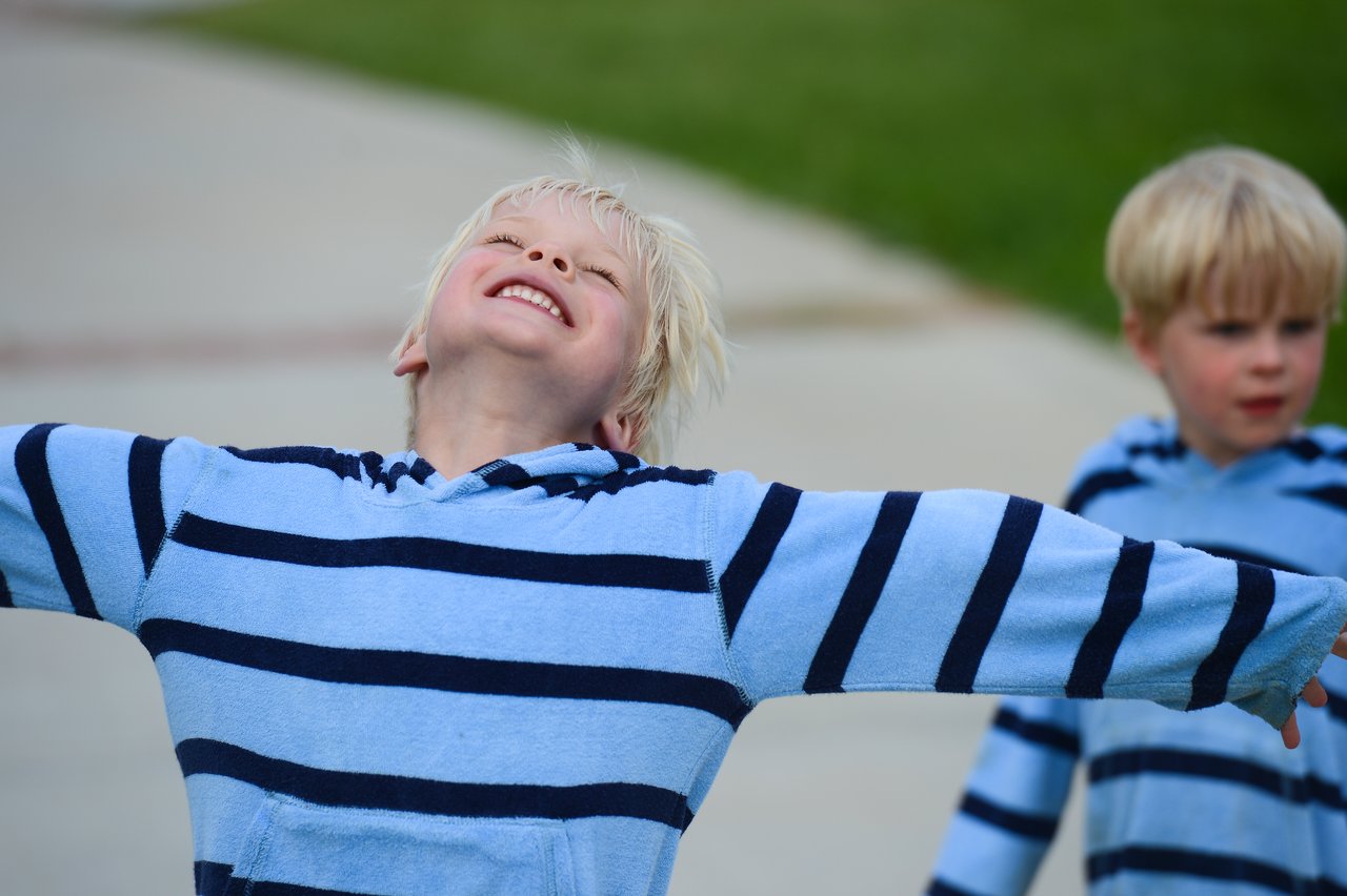 A smiling child in a striped hoodie spreads their arms wide, while another child watches in the background.