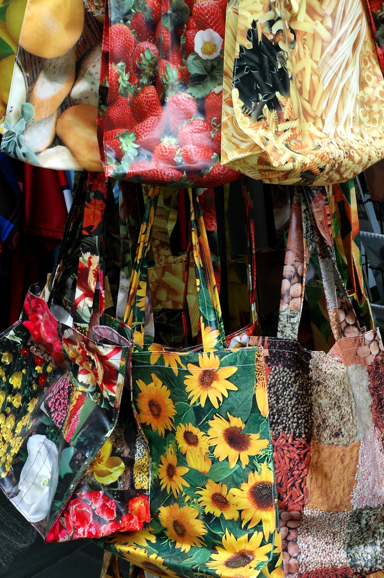 Colorful reusable bags with food and flower prints hang closely together on display.