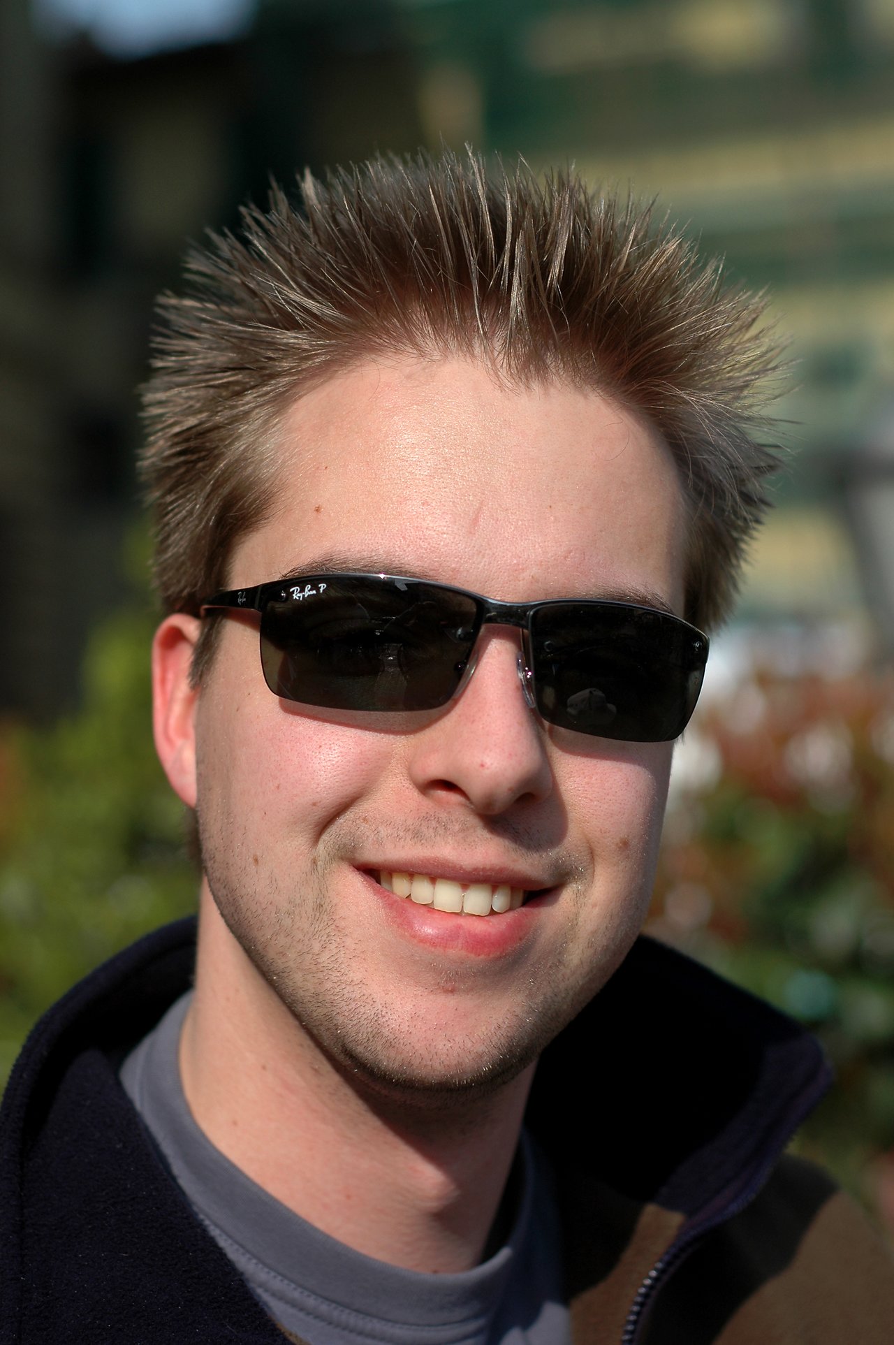A man with spiky hair and sunglasses smiles at the camera outdoors.