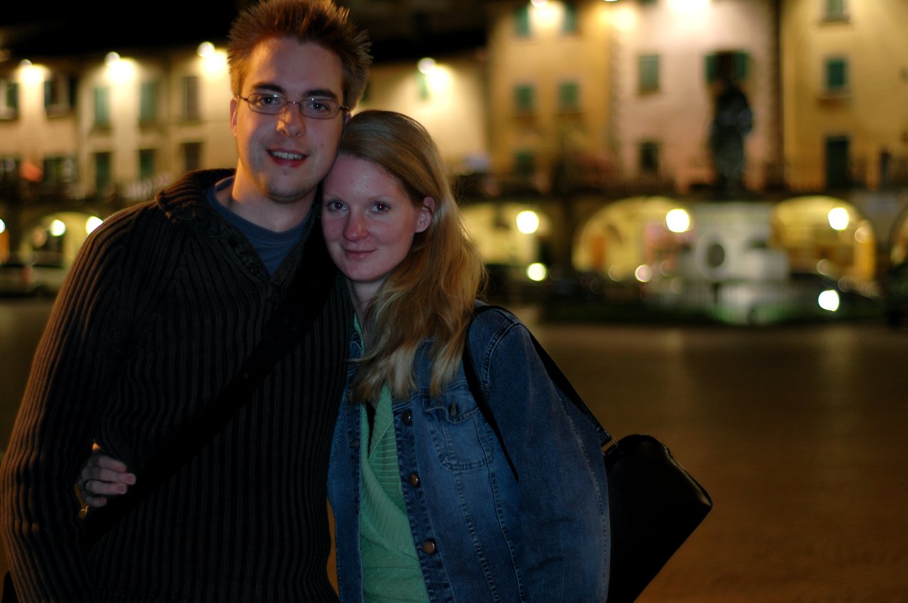 A young man and woman stand close together, smiling at the camera in a well-lit outdoor setting at night.