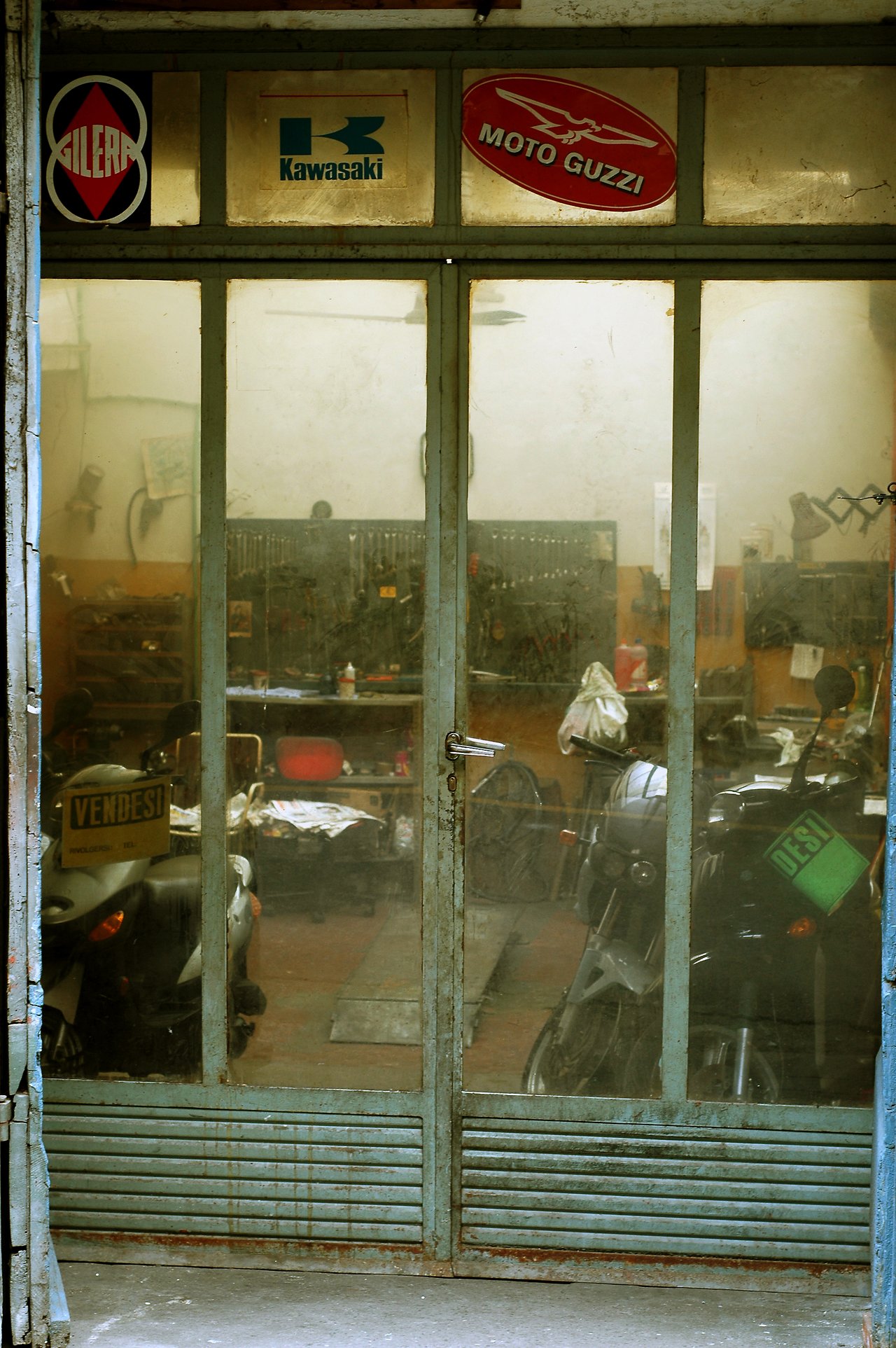 A dusty glass door reveals a cluttered garage with parked scooters, tools, and a "for sale" sign on one Vespa.