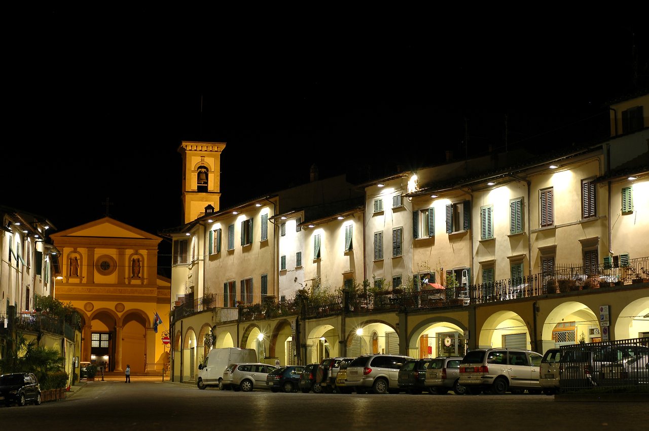 A well-lit triangular piazza in Greve in Chianti at night, featuring an old church and parked cars.