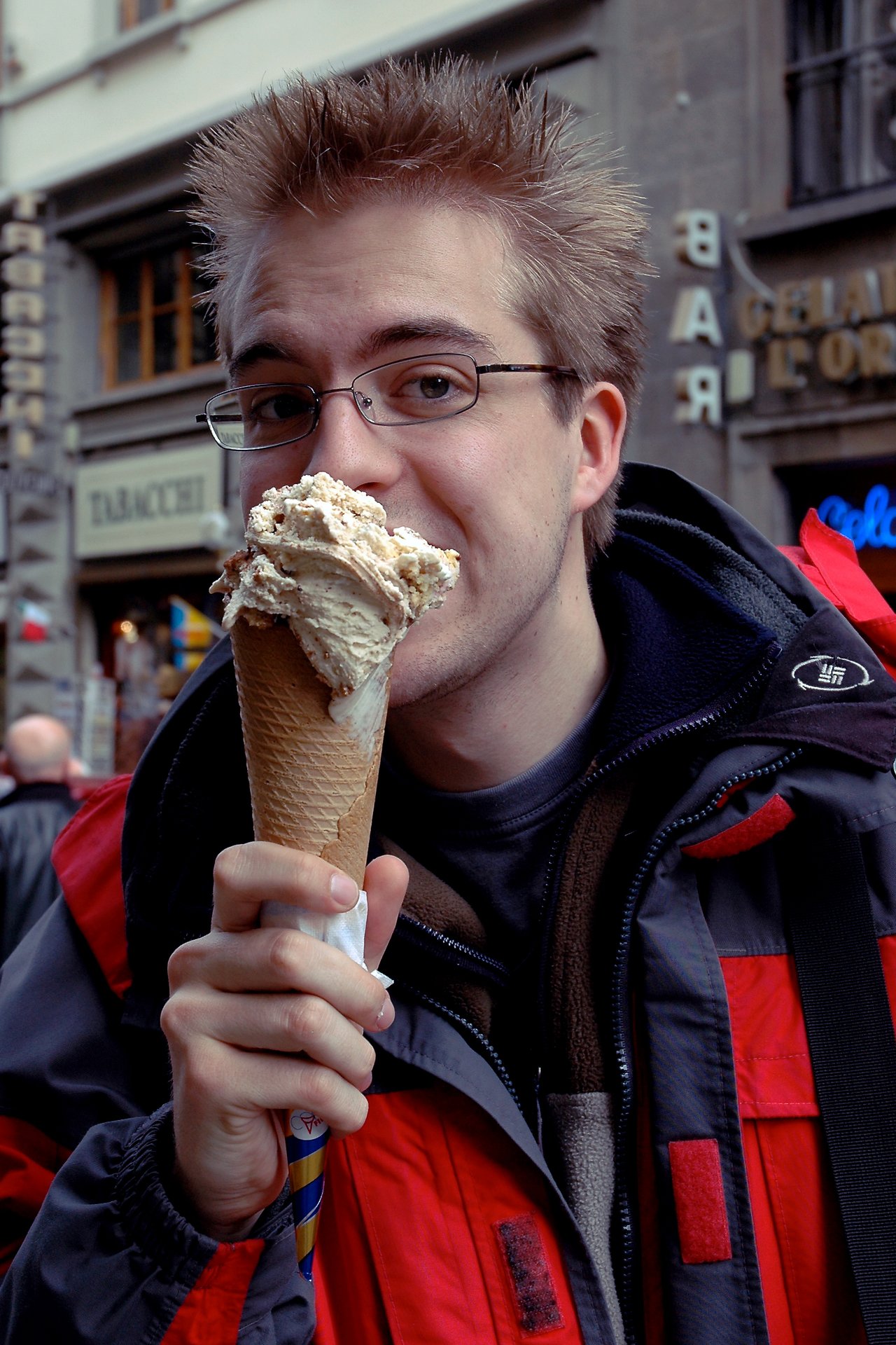 A person wearing glasses and a jacket enjoys a large ice cream cone while standing outside.