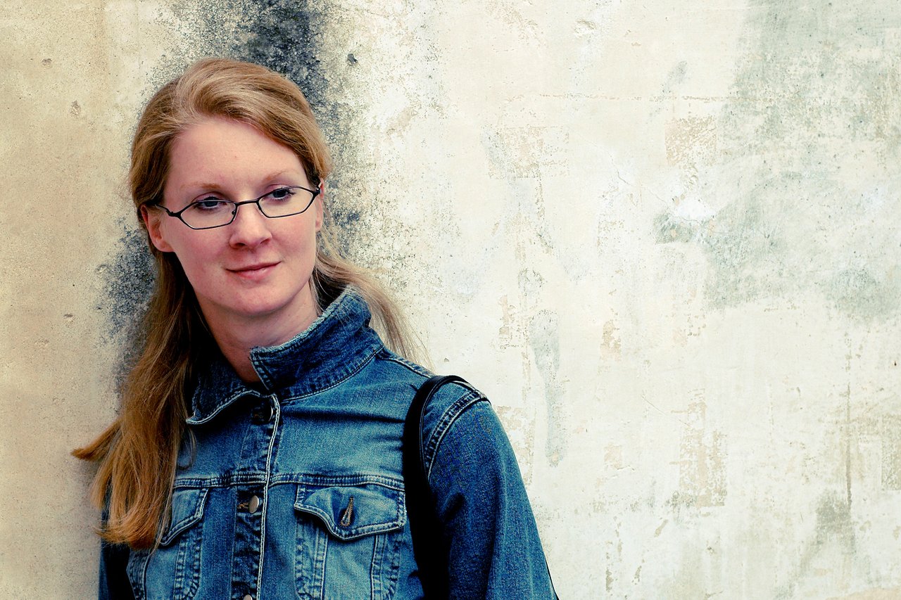 A woman with glasses and a denim jacket leans against a textured wall, looking slightly to the side.