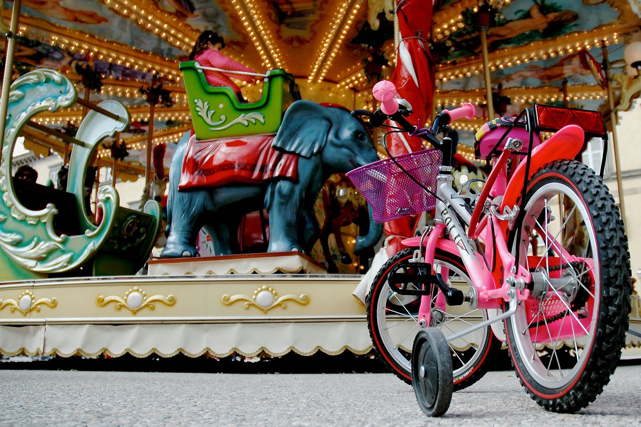 A pink children's bike with training wheels is parked in front of a carousel where a girl is riding.