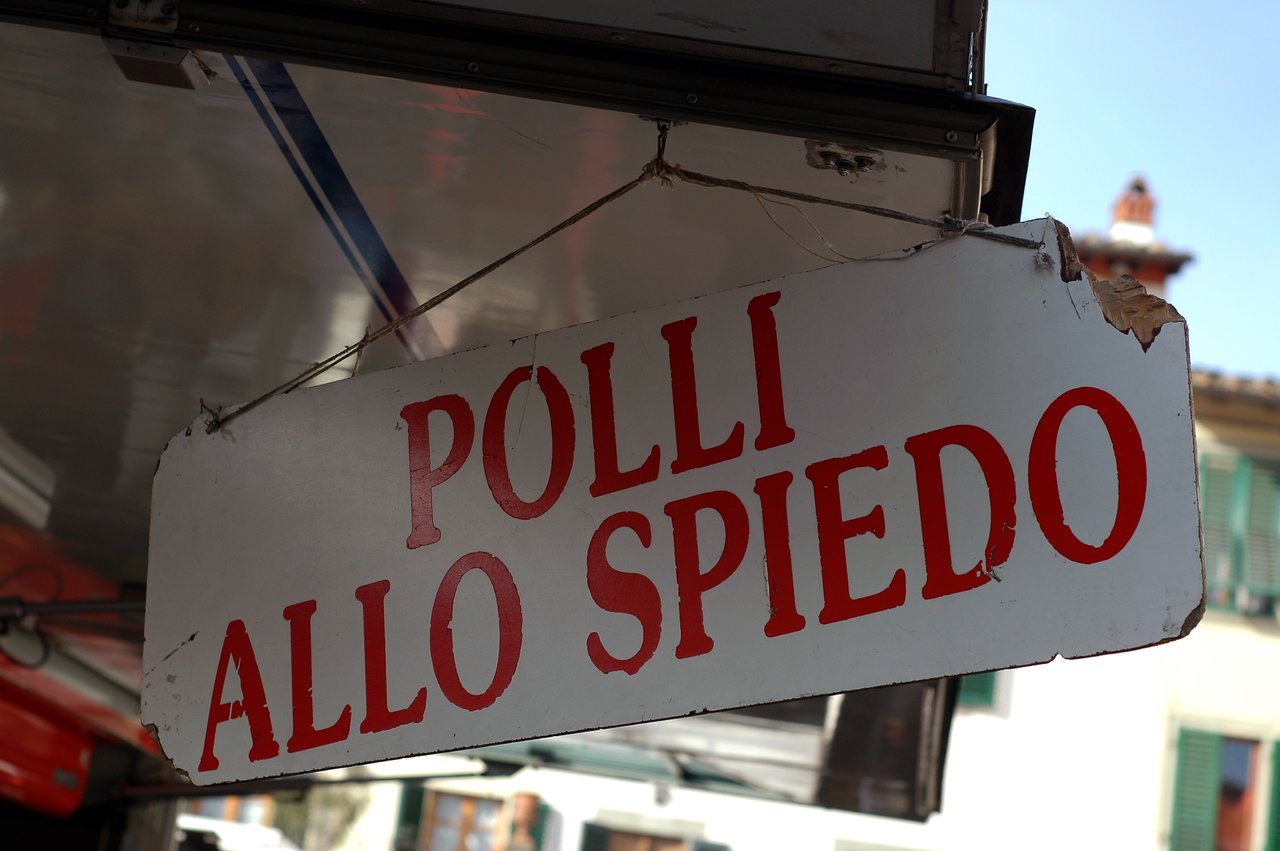 A worn white sign with red lettering advertises rotisserie chickens at a market stall in Greve in Chianti.