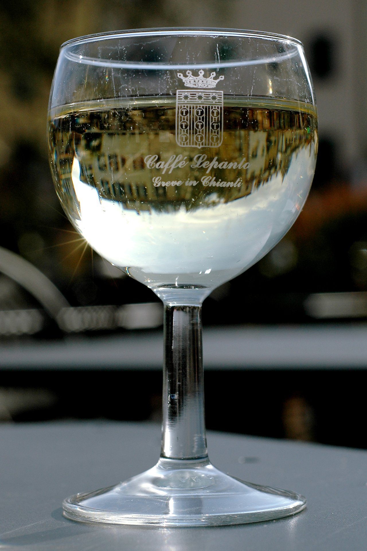 A glass of white wine on a table reflects an upside-down image of nearby buildings in its surface.