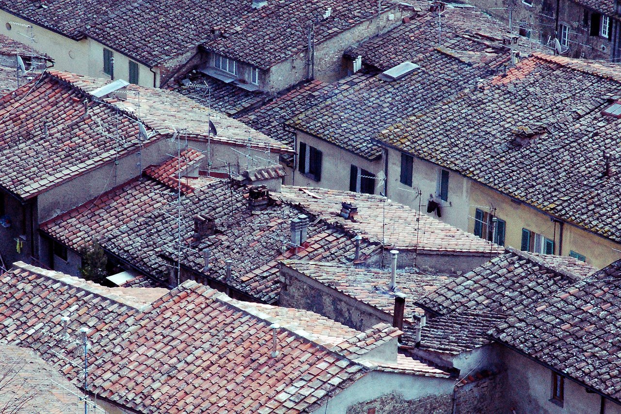 A view of old tiled rooftops in a Tuscan town, showing chimneys, antennas, and weathered surfaces.