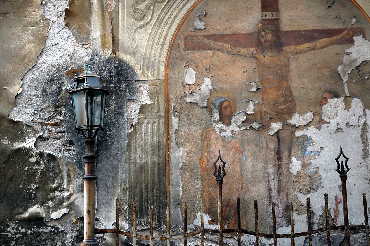 A faded fresco on a damaged wall depicts Jesus on the cross with two figures below, partially peeling away.
