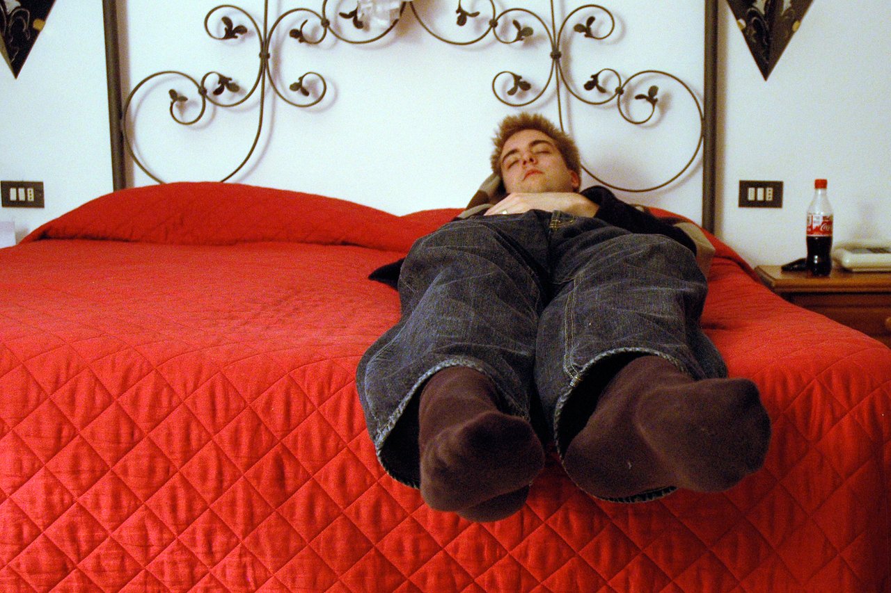 A man lies on a red bed with his feet hanging over the edge, showing the bed is too small.
