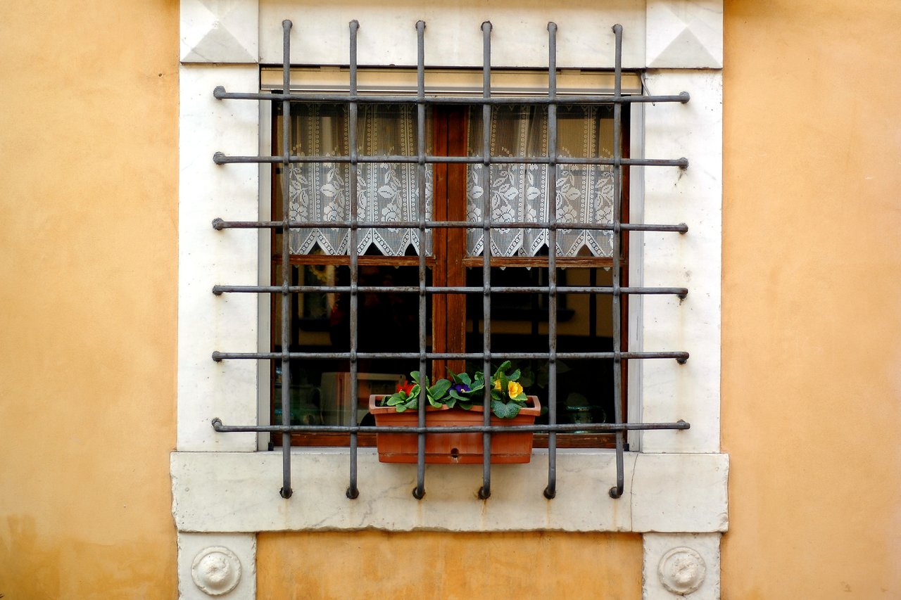A window with metal bars, lace curtains, and a flower box filled with colorful flowers on a beige wall.