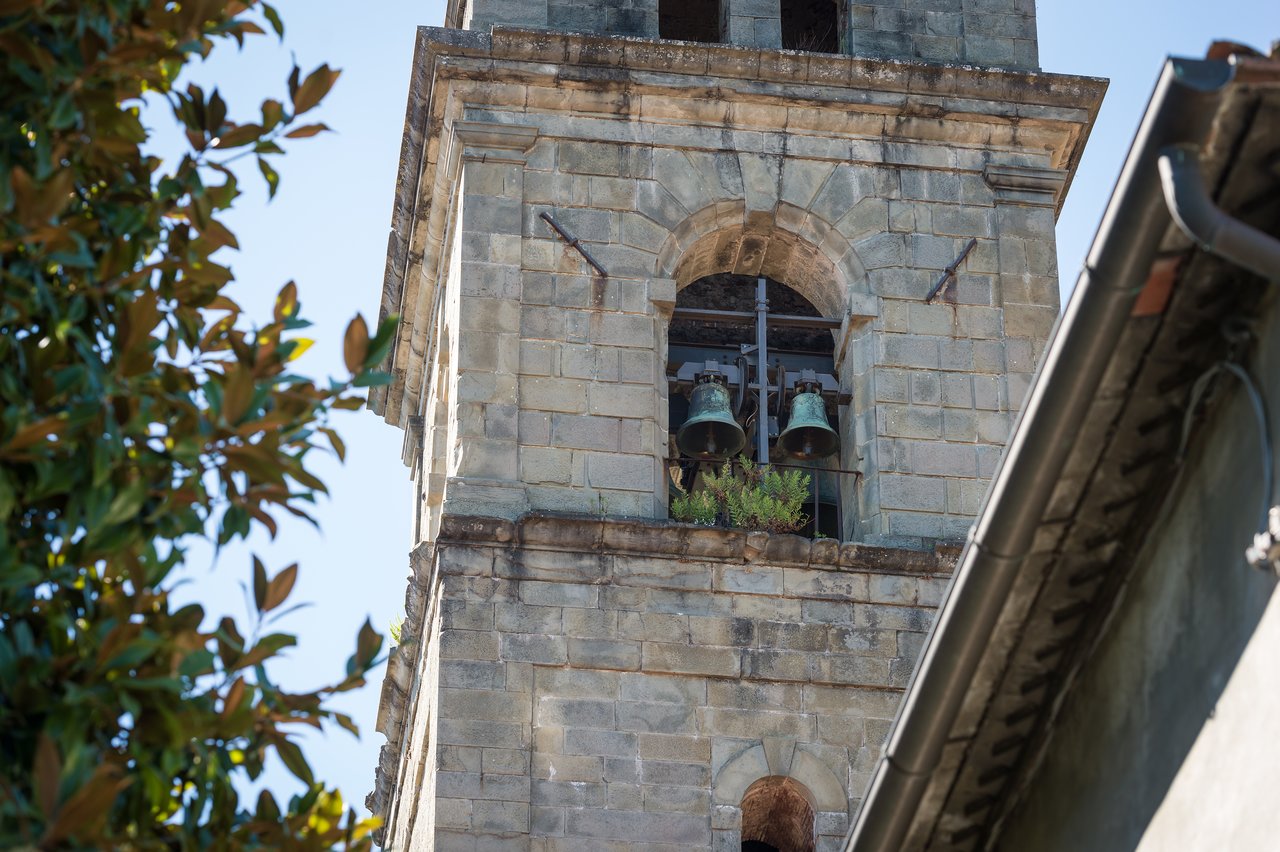 A stone bell tower with two large bells hanging inside an arched opening, surrounded by buildings and trees.
